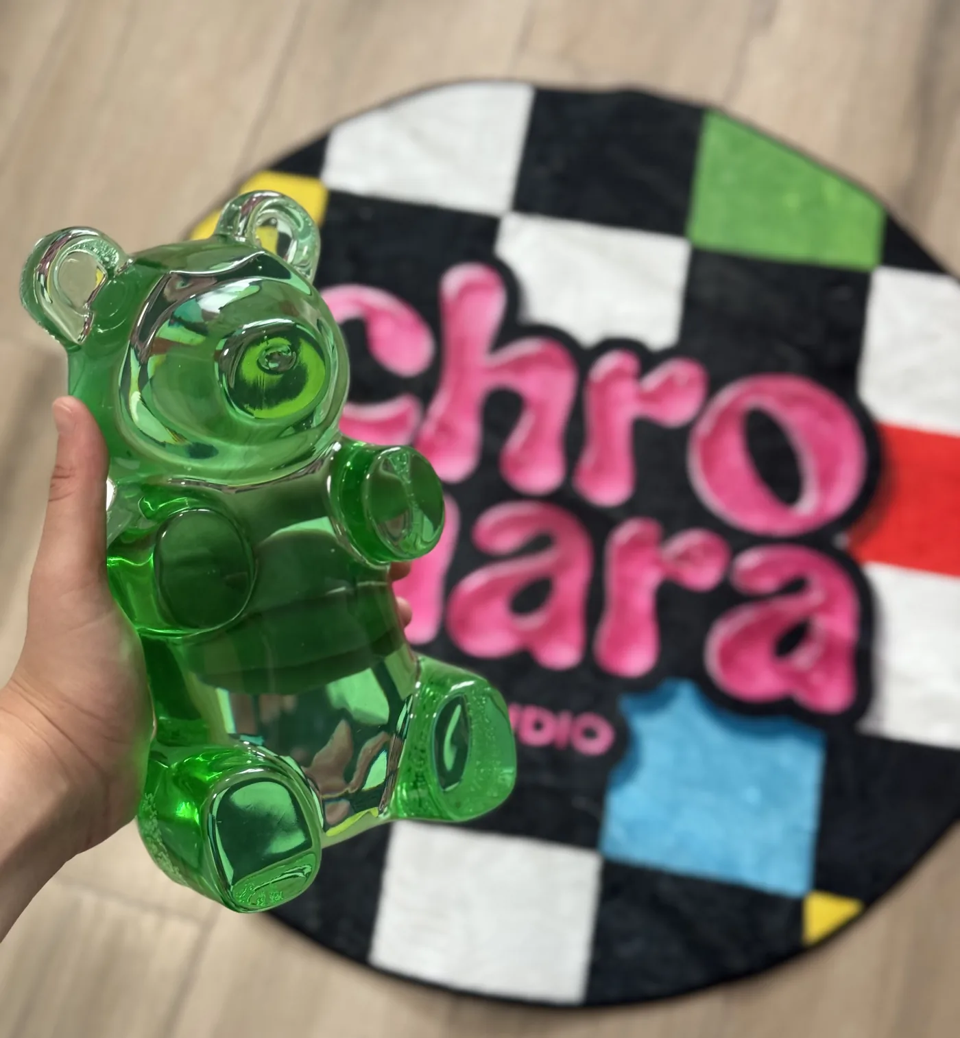 Green resin giant gummy bear held in a hand, on the Chromara Studio checkered rug