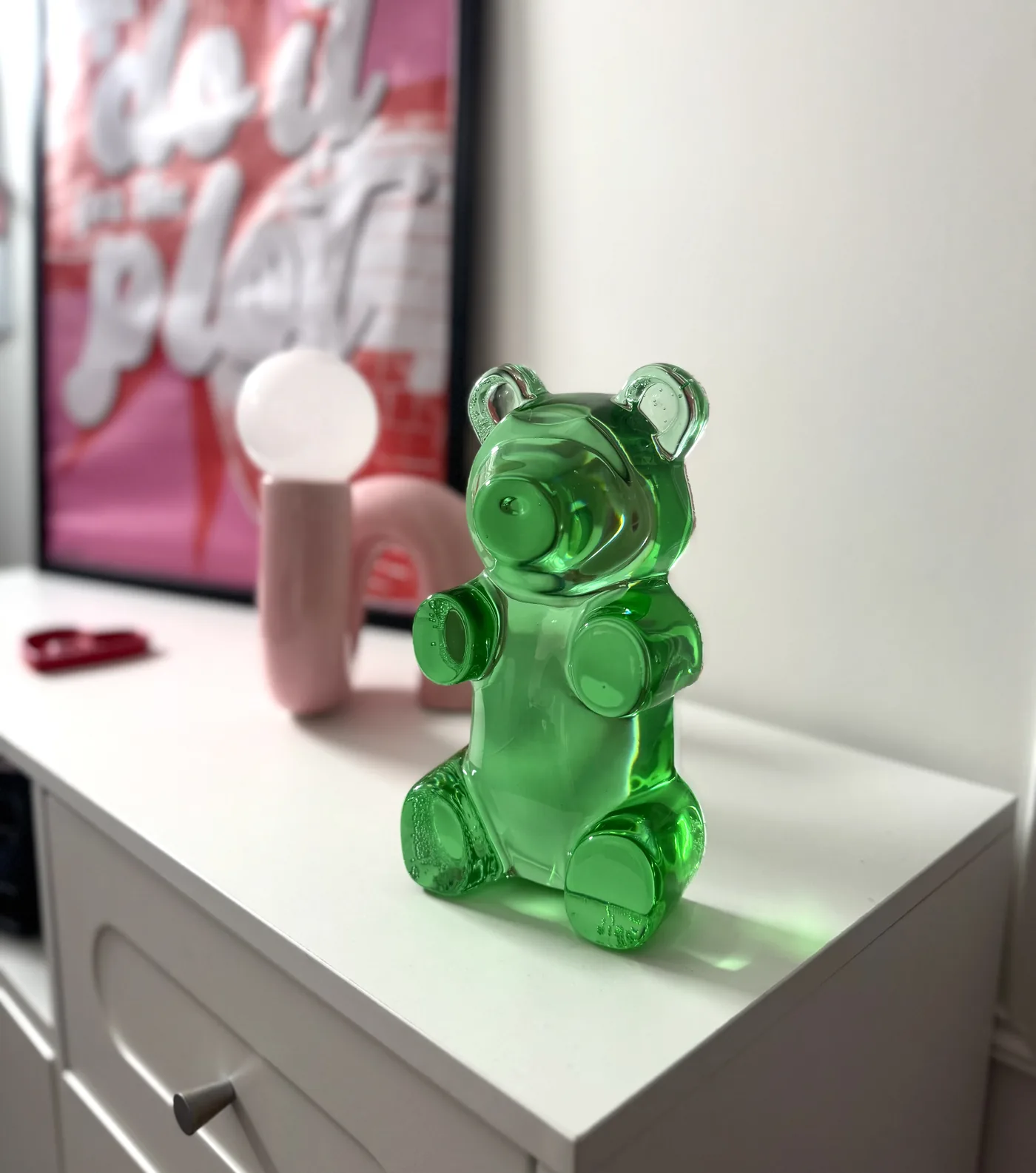 Grüner Gummibär aus Kunstharz auf einer weißen Kommode neben Lampe und Bild